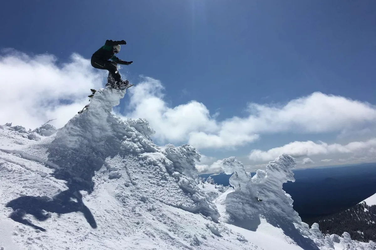 A snowboarder is captured mid-air against a stunning backdrop of snow-covered mountains and dramatic clouds. The image exudes energy and excitement, placing the viewer right in the heart of Central Oregon’s winter sports action.