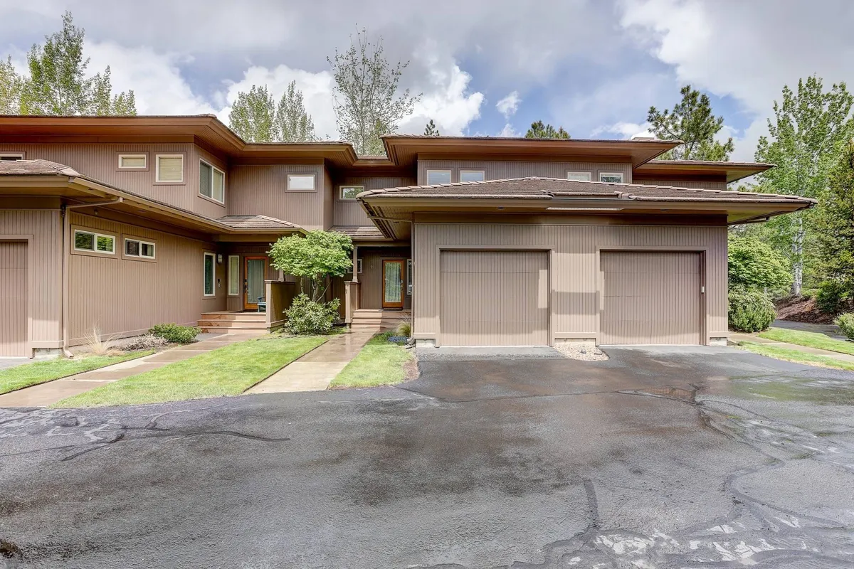 The image showcases the welcoming front entrance of a townhouse framed by lush green shrubbery, a manicured landscaped pathway, and the soft glow of sunset light. The spacious garage doors hint at convenience, while the inviting porch promises moments of relaxation and connection with nature.