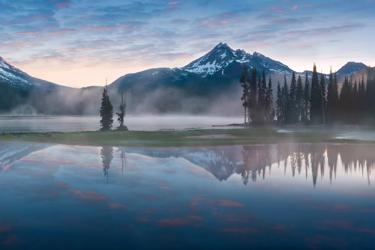 A breathtaking view of the Cascades mountains reflected in a serene lake, framed by lush trees and a light mist, evoking the dramatic natural backdrop described in the section.