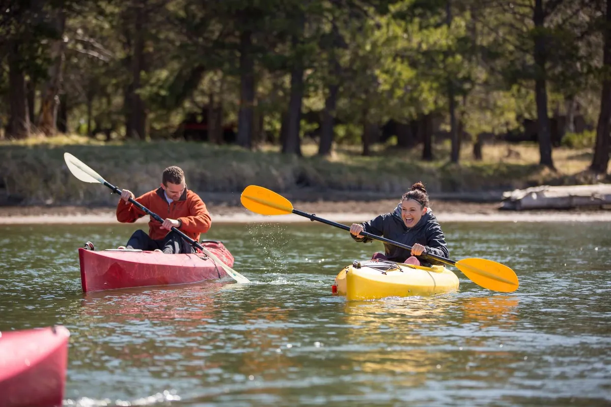 Participants paddle red and yellow kayaks across a serene water surface, framed by leafy trees in the background. The joyful expressions and vibrant colors evoke the thrill of water-based outdoor fun.