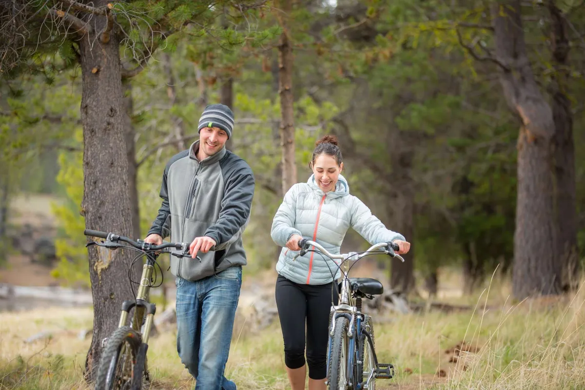 Two people are seen walking with bicycles along a natural landscape, surrounded by lush trees. Their relaxed and playful demeanor suggests the simple joys of exploring trails and experiencing the outdoors together.