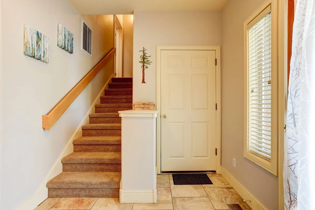 This image captures the entryway of a home, with a staircase, front door, decorative wall art, and a small tree decor. Natural light streams in, creating an inviting transition space that hints at welcoming guests or heading out for city adventures.