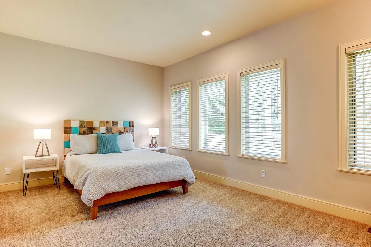 A bright bedroom featuring a bed with a colorful headboard, windows with blinds letting in natural light, and side tables with lamps, creating a peaceful morning or cozy night setting.
