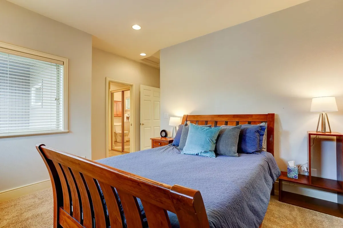 A serene bedroom with a wooden bed frame, decorative pillows, and a nightstand with a lamp. Natural light fills the room, emphasizing its soft linens and cozy, restful ambiance.